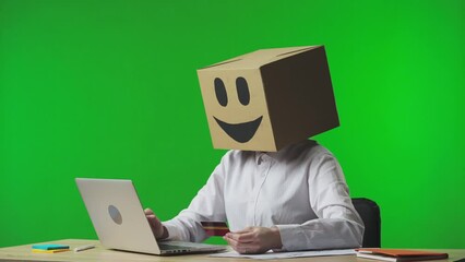 Woman in cardboard box with smiling emoji on studio green background. A worker makes a purchase online using a bank card and a laptop, gets frustrated with failure and turns the emoji to the sad side.