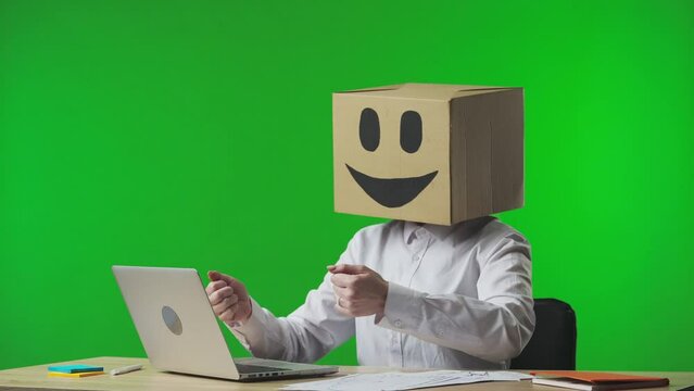 Woman in cardboard box with smiling emoji on her head on studio green background. The female worker is typing on a keyboard and showing a thumbs up gesture.