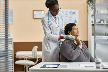 Mature short-haired Black woman patient feeling pain in neck while female doctor conducting medical...