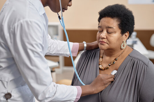 Medium close up shot of mature Black woman patient sitting with closed eyes in clinic while doctor conducting auscultation