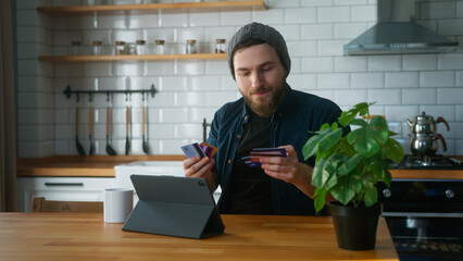 Anxious man with beanie sitting in kitchen at home takes his credit cards out of his pocket and...