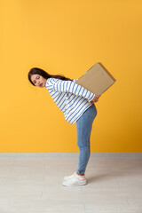 Pretty young woman carrying heavy parcel on her back against yellow background