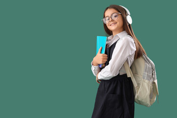 Little schoolgirl in headphones with copybook and backpack on green background