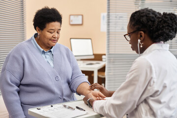 African American senior woman dressed in sweater sitting in clinic office with closed eyes while female doctor taking her pulse rate by hand - Powered by Adobe