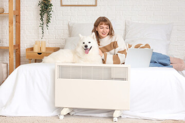 Young woman with laptop and Samoyed dog warming near radiator in bedroom