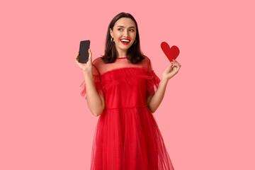 Happy young woman with mobile phone and paper heart on pink background. Online dating