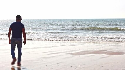 Mature man in a blue T-shirt, jeans, and cap, walking along the seashore with his back to the camera. In the background, the sea with waves, captured in slow motion. Conveys a sense of solitude.