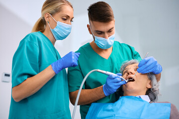 Two dentists performing dental procedure on senior woman's teeth.