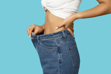 Young woman in loose jeans after weight loss on blue background, closeup