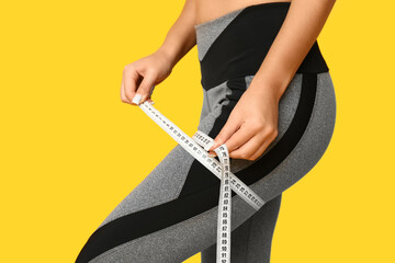 Young woman in sportswear measuring her thigh on yellow background, closeup. Weight loss concept - obrazy, fototapety, plakaty