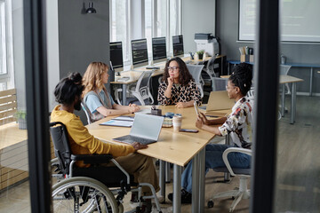 Three multi-ethnic women and indian man in wheelchair having business meeting at desk in office