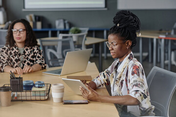 Obraz premium Black girl in smart casual outfit with dreadlocks typing on digital tablet sitting in office