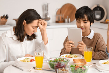 Teenage boy using tablet computer and his upset mother at table during dinner in kitchen. Family...