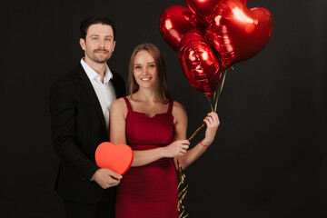 Beautiful romantic couple isolated on grey background. Attractive young woman in red dress and handsome man in suit are hugging with air balloons. Happy Saint Valentine's Day!