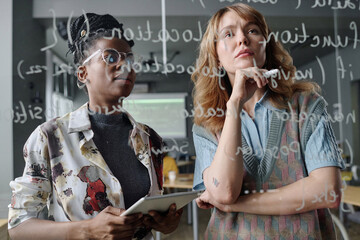 Two biracial female programmers writing code on glass board in modern IT company office