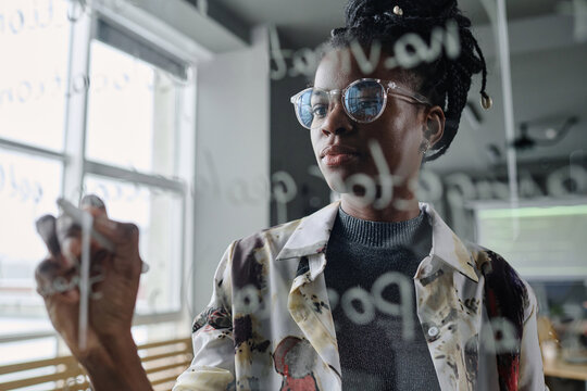 Young african american woman attentively writing program code with white marker on transparent wall