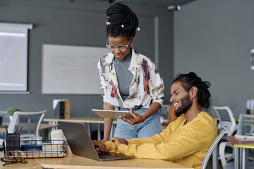 Cheerful indian programmer working on laptop at table in office, his black colleague standing nearby