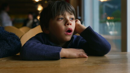 Bored small boy yawning while waiting at restaurant table by window. Child feeling boredom in...
