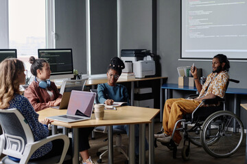 Indian coder in wheelchair showing presentation, his female coworkers listening and taking notes