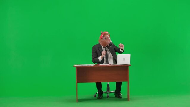 Man in business suit with horse head mask on studio green background. Businessman sitting at desk, listening to music with big red headphones and dancing. Concept of heavy office work.