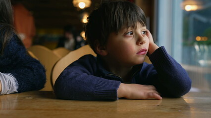 Bored small boy yawning while waiting at restaurant table by window. Child feeling boredom in...
