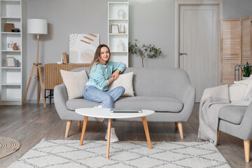 Fototapeta premium Young woman sitting on grey couch in living room