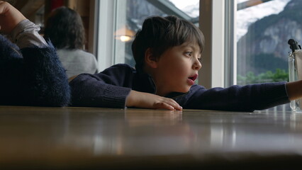 One small boy at restaurant diner waiting for food to arrive. Child leaning on table by window...