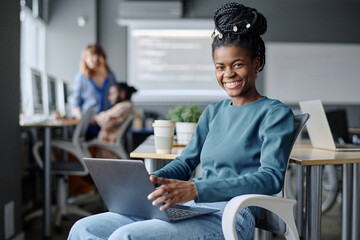 Young female african american coder sitting on chair with laptop on her lap smiling at camera