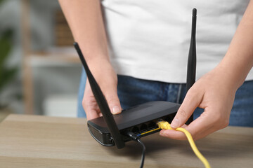 Woman inserting ethernet cable into Wi-Fi router at table indoors, closeup - obrazy, fototapety, plakaty