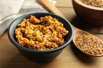 Whole grain mustard in bowl and dry seeds on wooden table, closeup