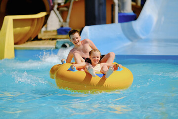 Boy and girl have fun on water slide in outdoor aquapark. Little children floating on yellow...