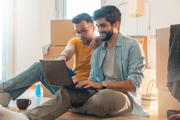 Handsome young men sitting on floor with cardboard boxes around, using laptop, making online...