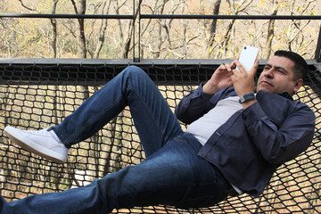 Dark-skinned Latino adult man uses his cell phone to work and make video calls over a network while relaxing in his forest cabin