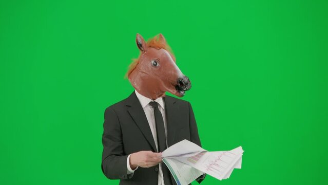 A man in a business suit with a horse head mask on a green studio background. A businessman is looking through documants and dancing merrily. Hard office work concept.