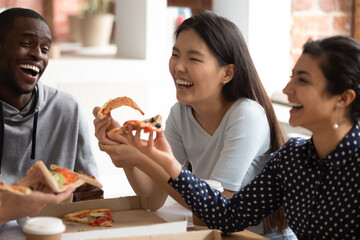 Smiling multiethnic young people have fun eating tasty Italian fast food from takeaway delivery, overjoyed multiracial students laugh joke enjoying delicious pizza on lunch break in class together - obrazy, fototapety, plakaty