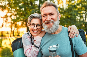 Portrait of active sincere carefree smiling happy modern gray-haired couple shouldering bag with sports mats, man holding water. Sports jogging, training in the park in the morning. - Powered by Adobe