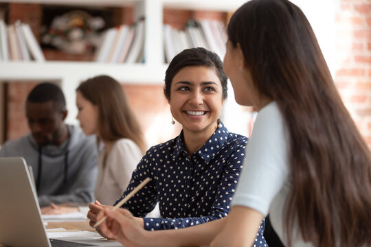 Happy indian female student talk discuss sharing ideas brainstorming at lesson with teammate, smiling ethnic girl cooperating work on school project using laptop studying in classroom together