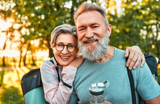Portrait of active sincere carefree smiling happy modern gray-haired couple shouldering bag with sports mats, man holding water. Sports jogging, training in the park in the morning.