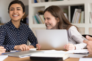 Excited multiethnic girls sit at desk in classroom laugh brainstorming learning preparing for exam, happy smiling multiracial female students have fun gather work on shared project or study together