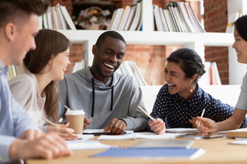 Smiling international young people sit at desk have fun studying preparing group project together, happy overjoyed multiethnic students laugh joke working in team cooperating or brainstorming in class