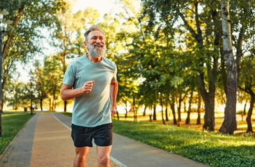 Physical activity on retirement. Happy aged man in sport clothes running alone at green summer park...