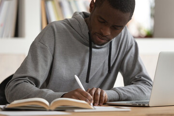 Concentrated african American millennial man sit at desk studying using laptop making notes in notebook, focused smart biracial male student writing reading textbook learning, education concept - Powered by Adobe