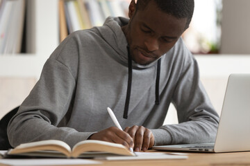 Concentrated african American millennial man sit at desk studying using laptop making notes in notebook, focused smart biracial male student writing reading textbook learning, education concept