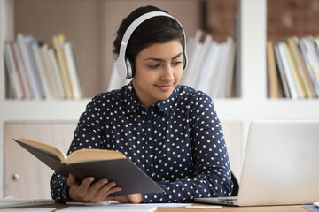 Smiling millennial indian girl in headphones read textbook study watching webinar on laptop, happy...