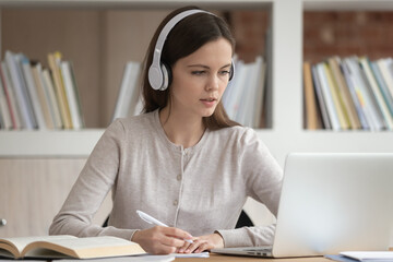 Focused millennial girl wearing modern Bluetooth earphones look at laptop screen make notes in...