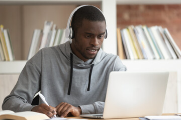 Concentrated biracial male student in Bluetooth headphones look at laptop make notes taking online...