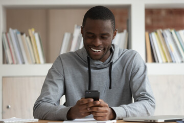 Smiling african American male student sit at desk browsing wireless internet on smartphone, happy...
