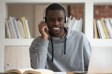 Smiling african American millennial guy in Bluetooth headphones look at laptop screen watching...