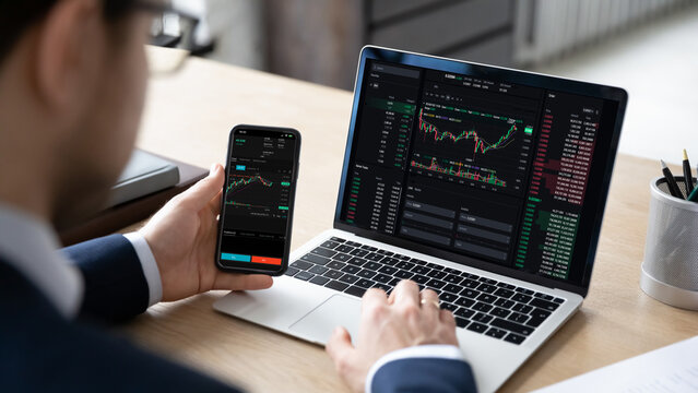 Male trader sits at desk hold smart phone with stock market data, using laptop display graph and chart, analyzing investment, check trading stocks information, view over shoulder. Growth, profit, apps