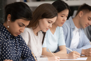 Concentrated multiracial young people sit in row at table busy writing examination or test in...